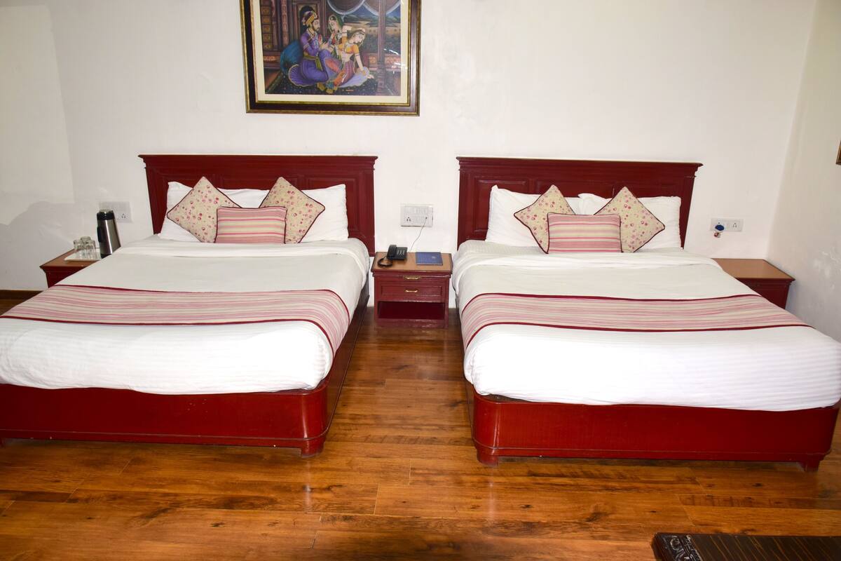 A hotel room with two beds, wooden headboards, a painting on the wall, and a small bedside table with a phone and a book.