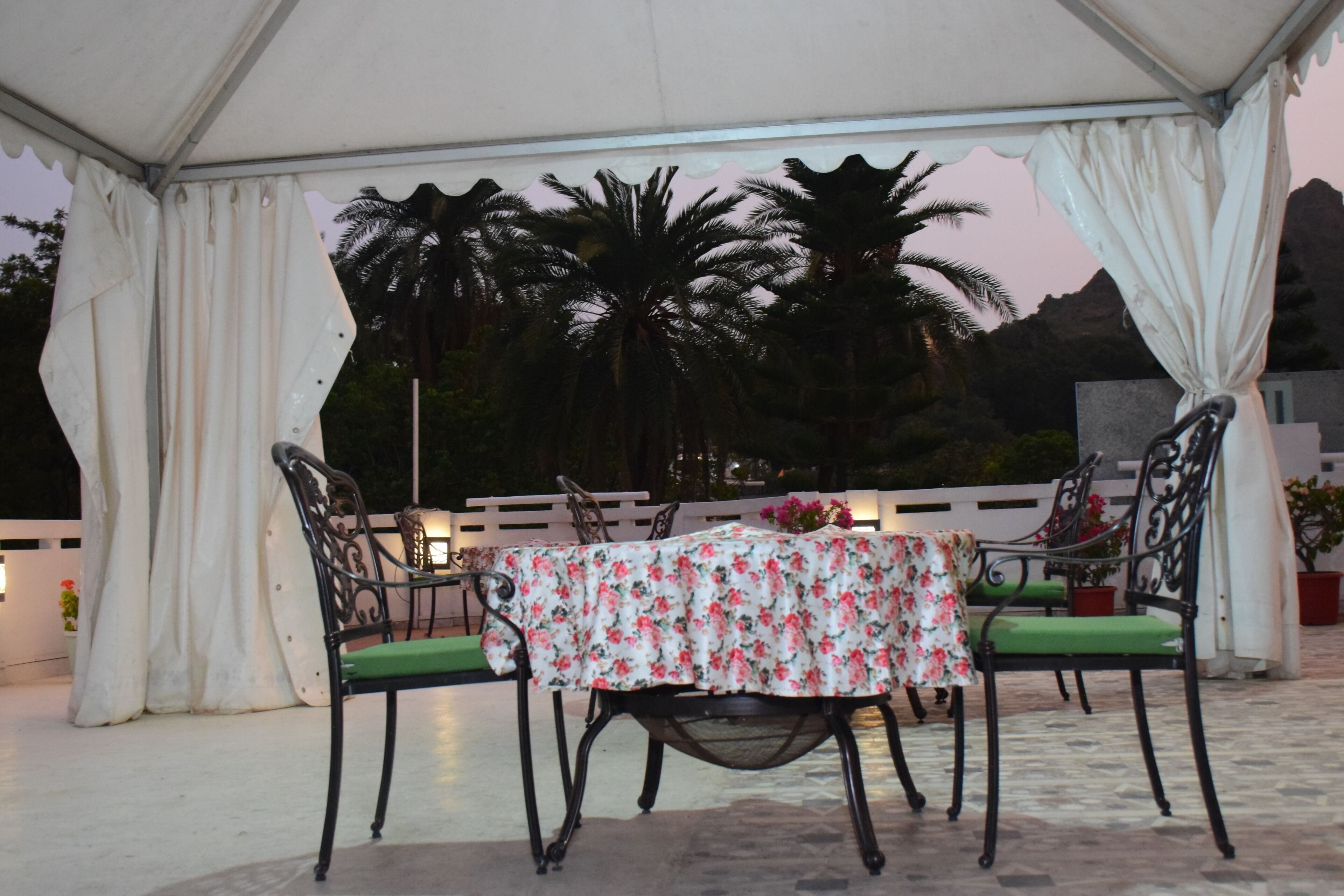 A covered outdoor seating area with a table and chairs, surrounded by curtains and palm trees.