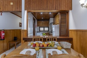 Un comedor con mesa de madera dispuesta para el desayuno, con frutas y jugo, y vistas a la cocina.