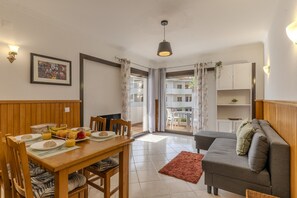 Un comedor con una mesa de madera, sillas y una disposición para el desayuno. En la sala de estar, hay un sofá gris y una alfombra roja.
