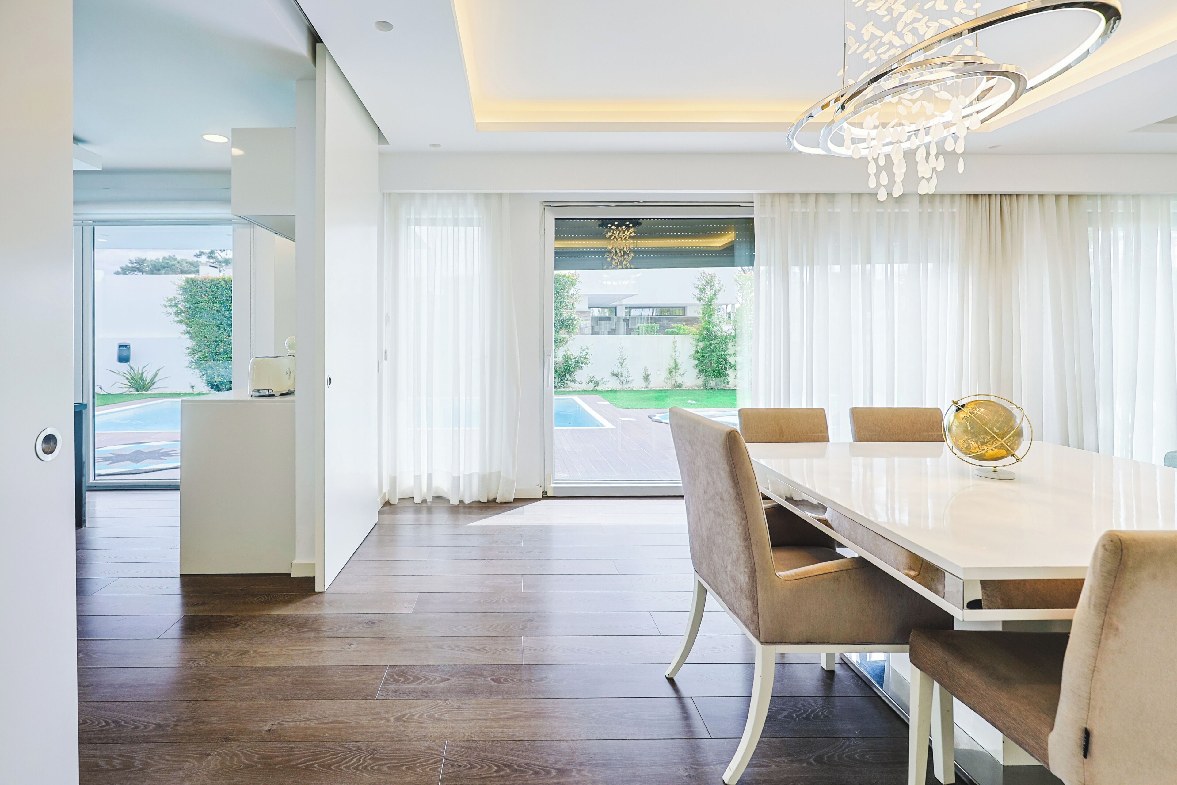 A modern dining room with a white table, beige chairs, and a chandelier.