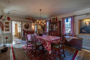 A dining room with a red tablecloth, wooden chairs, a chandelier, and a large painting on the wall. - Beautiful peaceful country home on 18 acres built 1800 (Ryegate)