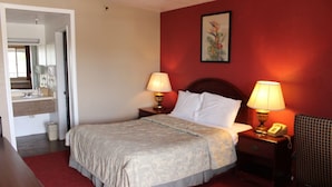 A hotel room with a bed, two bedside lamps, a nightstand with a telephone, a mirror, and a red wall with a framed picture. - Days Inn by Wyndham Victorville/hesperia I-15 & P (Victorville)