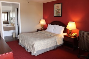 A hotel room with a bed, two bedside lamps, a nightstand with a telephone, a mirror, and a red wall with a framed picture.