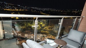 Ein Balkon mit Glastrennwand, zwei graue Polsterstühle, ein runder Tisch und Blick auf eine beleuchtete Stadtlandschaft bei Nacht.