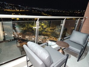 Ein Balkon mit Glastrennwand, zwei graue Polsterstühle, ein runder Tisch und Blick auf eine beleuchtete Stadtlandschaft bei Nacht.