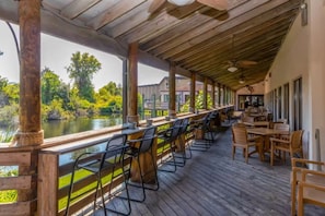 Un área de bar al aire libre con techo, suelo de madera, sillas y mesas de metal, con vistas a una masa de agua y vegetación.
