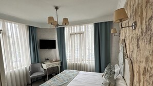 A bedroom with a bed, a desk, a chair, a TV, and a window with curtains. - Port Hotel (Istanbul)