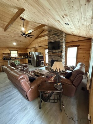 A log cabin interior with a leather sofa, a coffee table, a stone fireplace, and a kitchen area. - Bigfoot's Crossing is an authentic log cabin about 10 minutes from Charleston. (South Charleston)