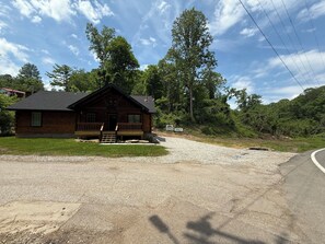 A wooden cabin with a porch, surrounded by trees and a gravel driveway.