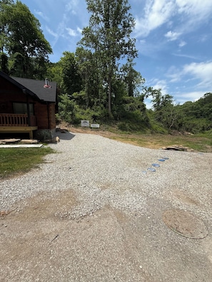 A gravel driveway leading to a wooden cabin with a chimney, surrounded by dense trees and a clear sky.