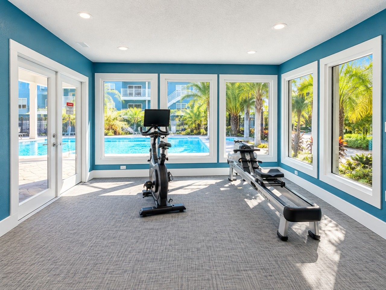 A gym with exercise equipment and large windows overlooking a pool area.