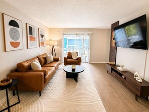 A living room with a brown leather sofa, a round coffee table, a TV on a wooden stand, and a large window with blinds. - Stylish Tampa Getaway | Airport, Stadiums & Beaches (Tampa)
