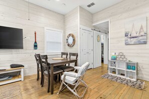 A room with a wooden floor, a dining table with chairs, a TV, a white door, and a wall with a painting. - Coastal Command | Beach Bungalow | 11th st (Port Aransas)