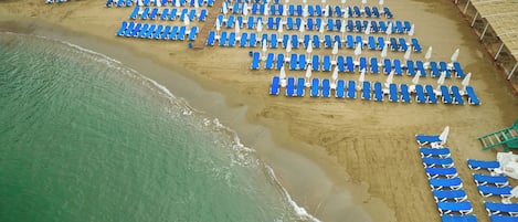 An aerial view of a beach with rows of blue lounge chairs and white umbrellas, a sandy shore, and a playground in the background.