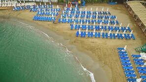 Vista aérea de uma praia com fileiras de cadeiras-de-sol azuis e guarda-sóis brancos, uma orla arenosa e um parquinho ao fundo.