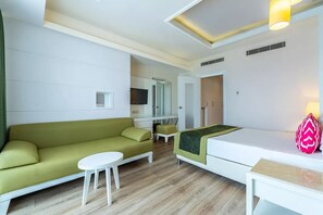 Quarto de hotel moderno com sofá verde, cama branca, mesinha pequena branca e televisão fixada na parede.