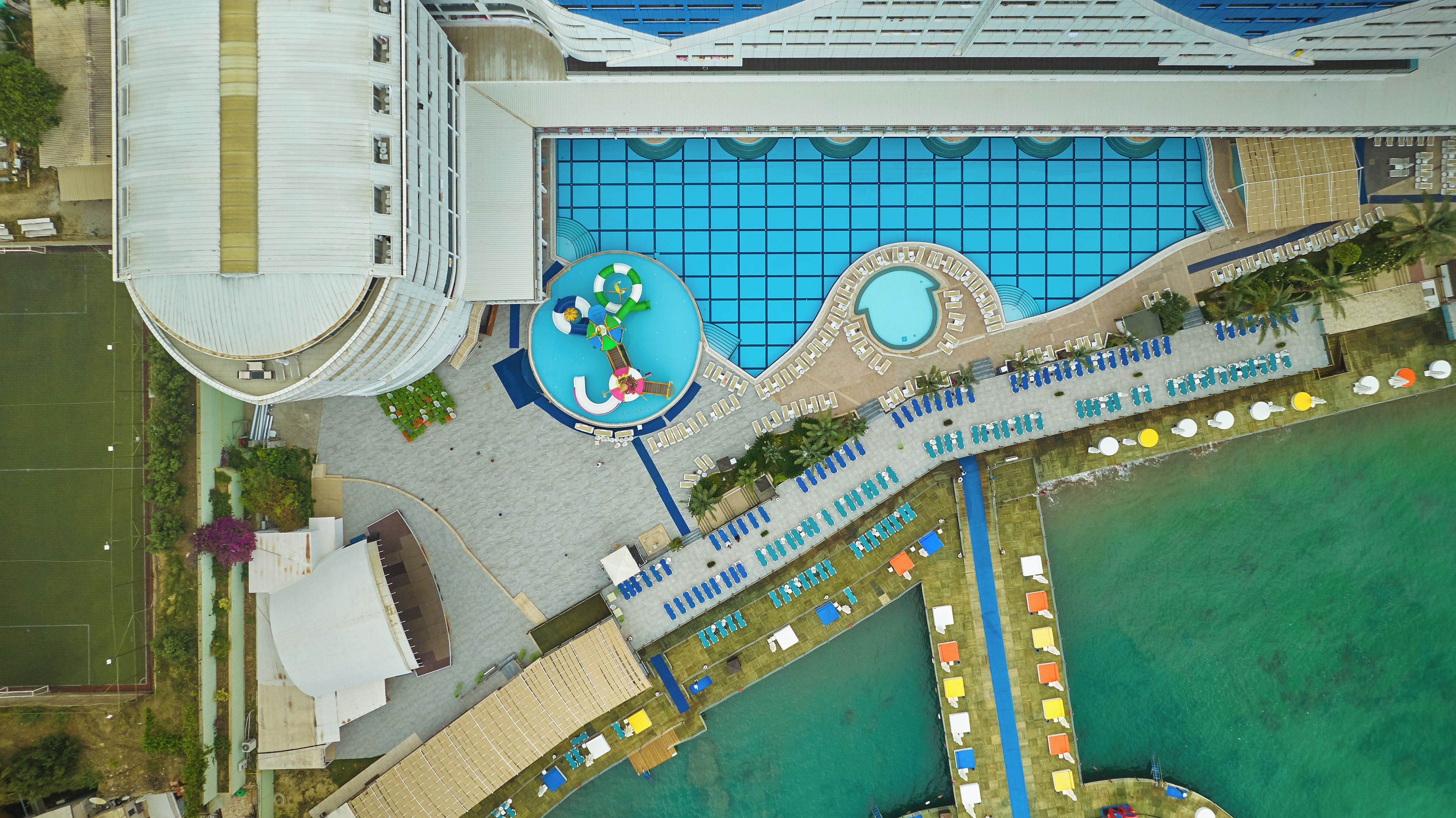 An aerial view of a hotel with a large swimming pool, multiple lounge areas, and a tennis court.