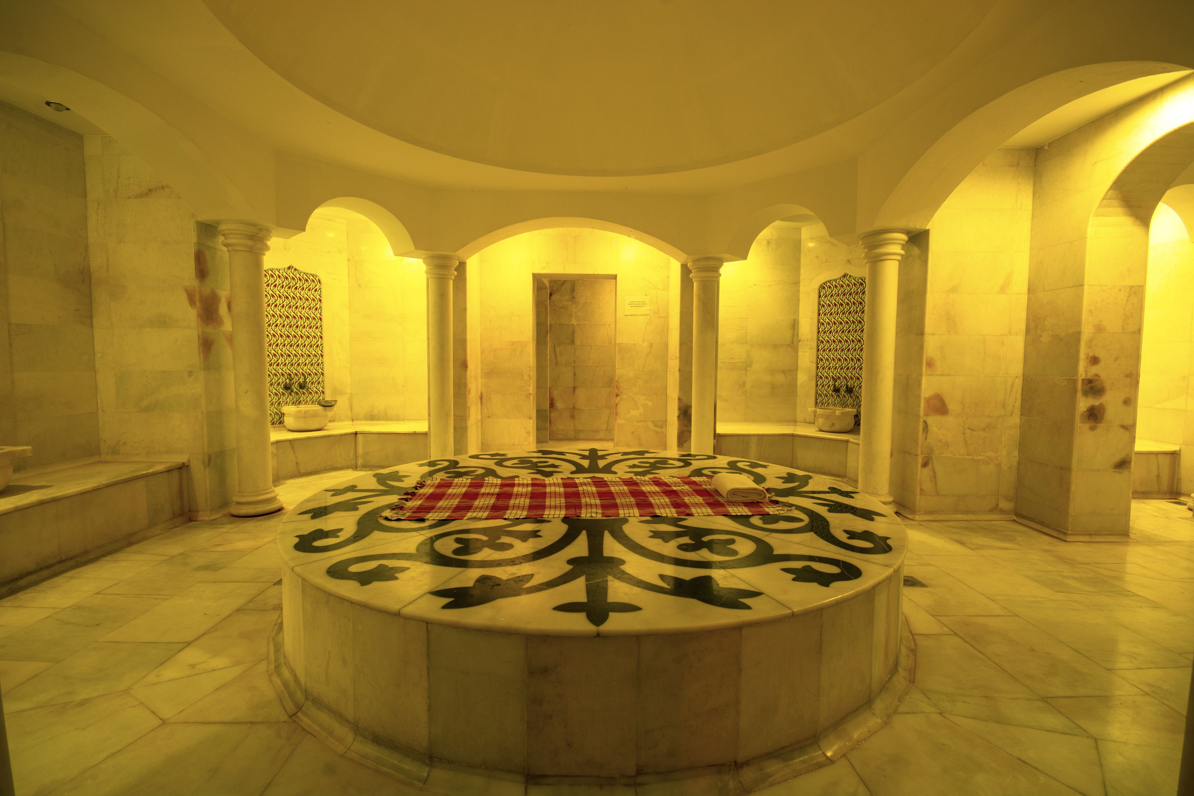 A circular marble fountain with a checkered cloth in the center, surrounded by arches and columns in a well-lit interior space.