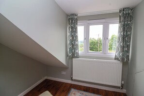 Ein Zimmer mit schrägverdeckter Decke, einem Fenster mit gemusterten Vorhängen, einem weißen Heizkörper und einem Holzboden.