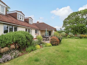 Ein zweigeschossiges Haus mit Ziegeldach, einer Terrasse mit Gartenmöbeln und einem gepflegten Garten mit verschiedenen Pflanzen und einem Rasen.