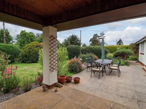 Eine überdachte Terrasse mit Tisch und Stühlen, umgeben von Kübelpflanzen und einem gepflegten Garten.