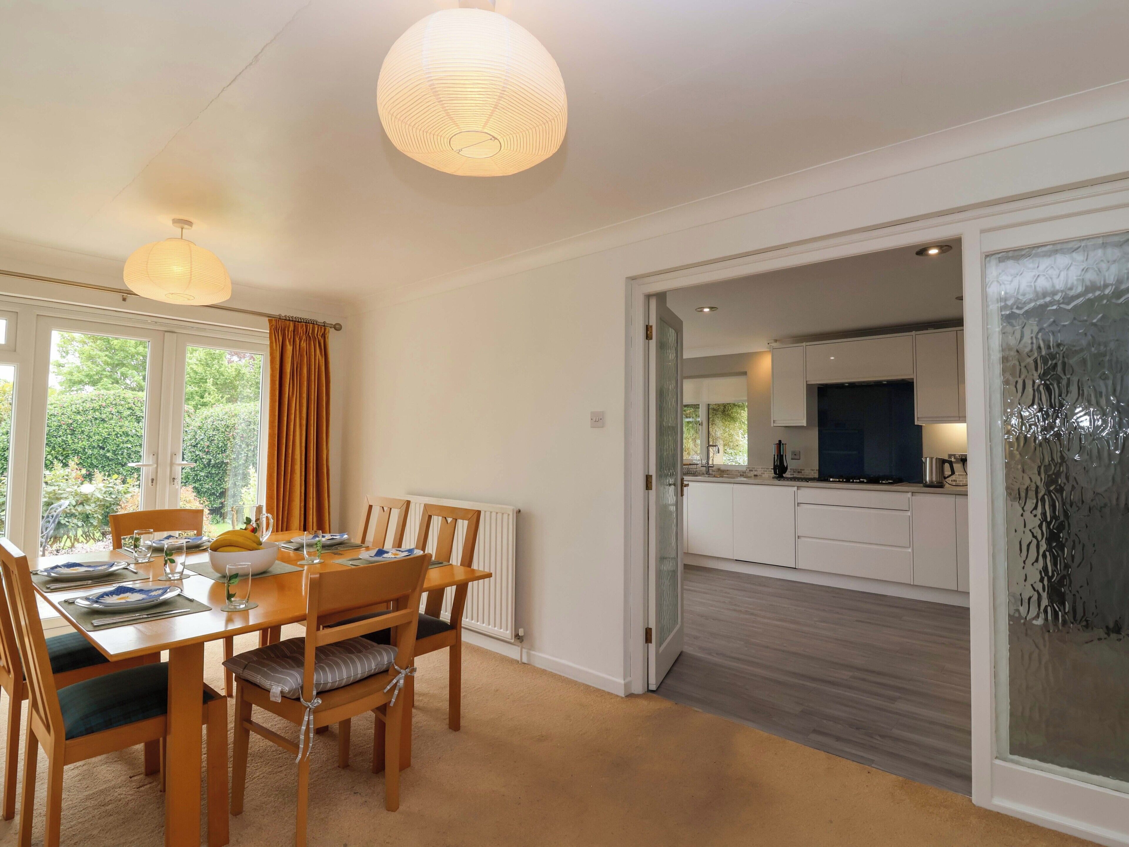 A dining area with a table set for four, a kitchen in the background, and a view of greenery outside.