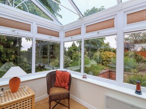 A conservatory with a chair and a small table, large windows, and a view of a garden. - Meadowcroft (Watchet)
