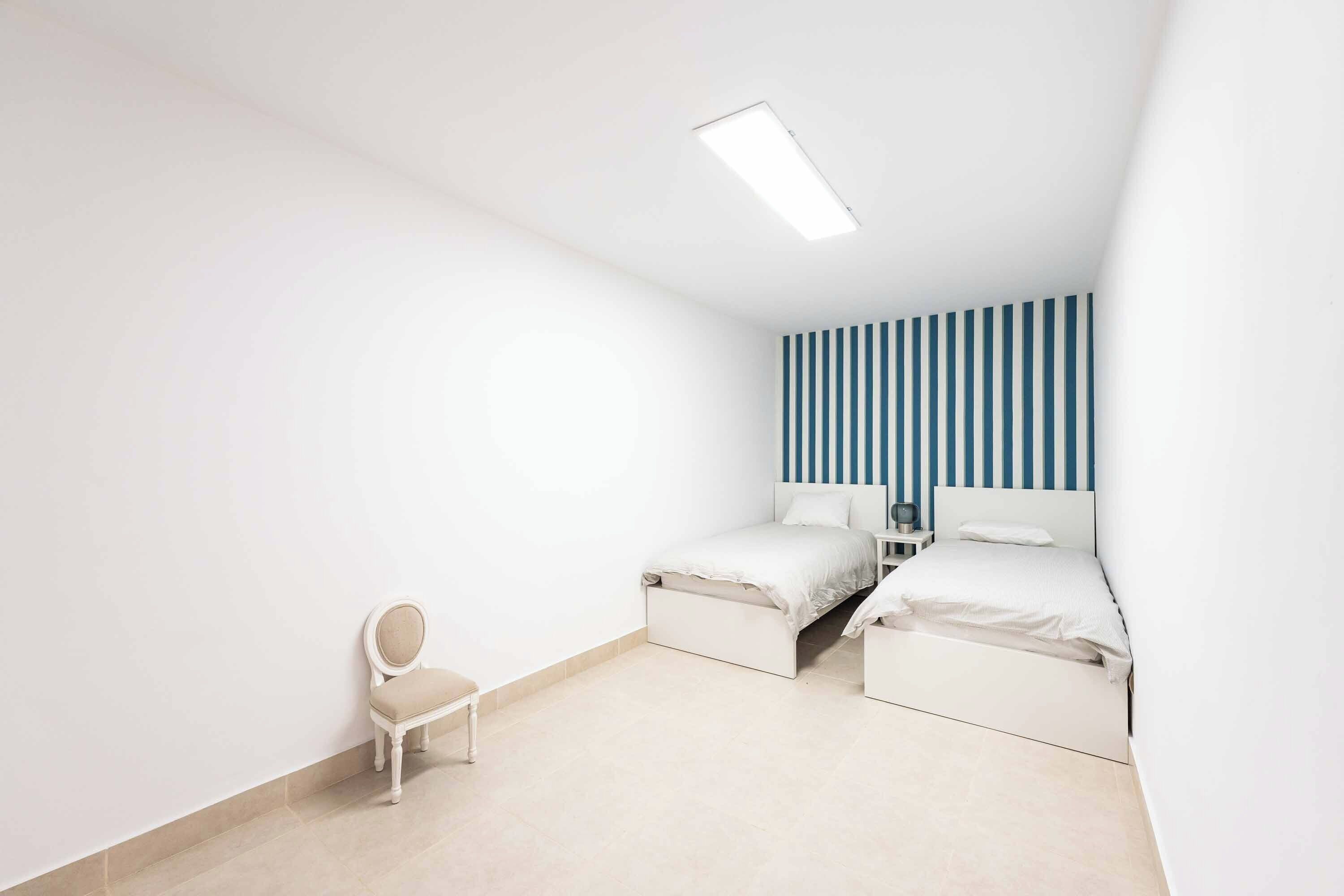 A bedroom with two beds, a chair, a bedside table, and a striped wall.
