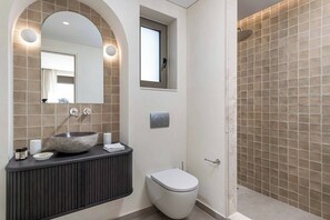 Ein Badezimmer mit einem Becken aus Stein, Toilette, Dusche und einem kleinen Fenster.