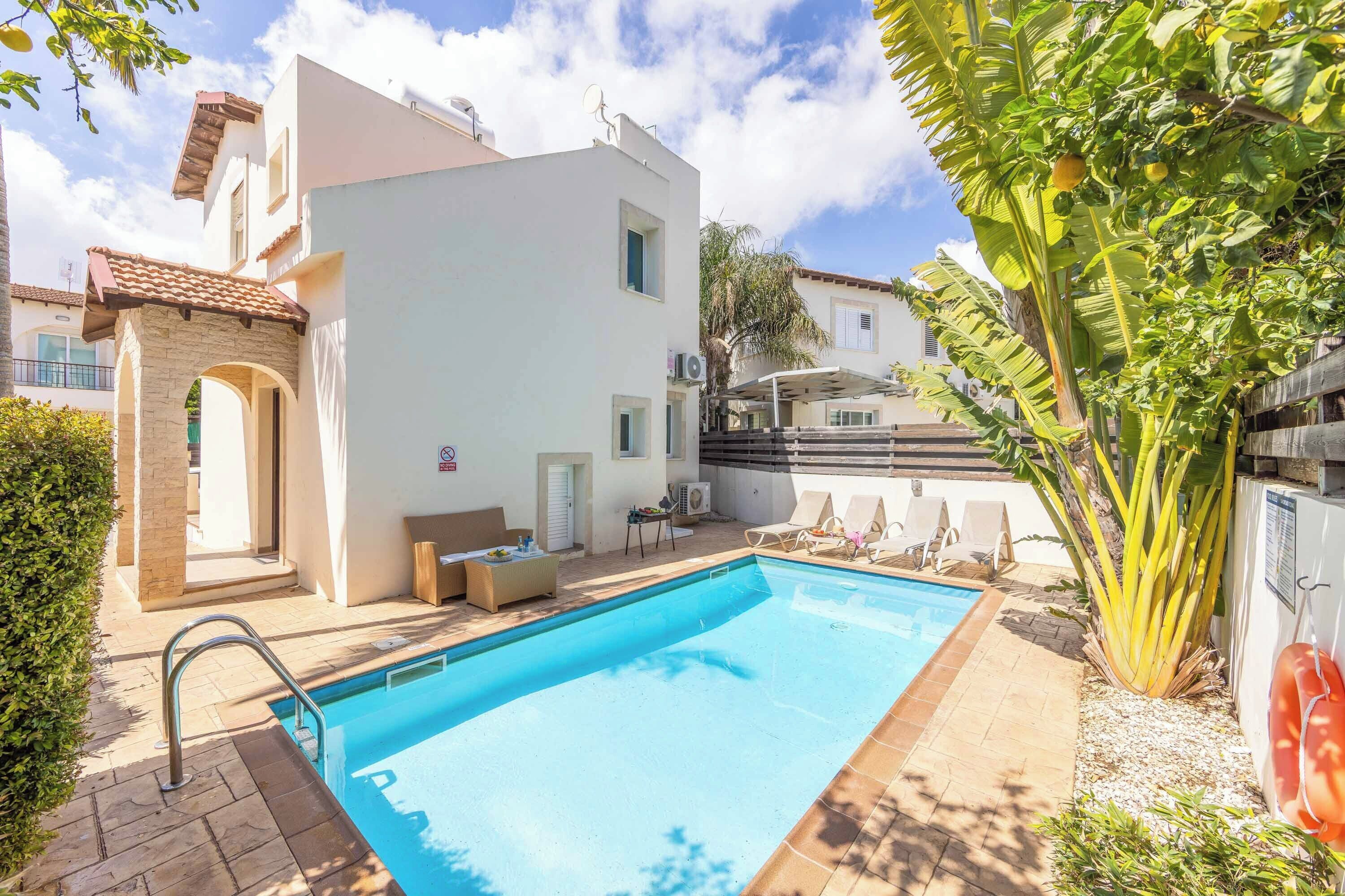 A swimming pool with a patio area, a white building, and a large banana tree.