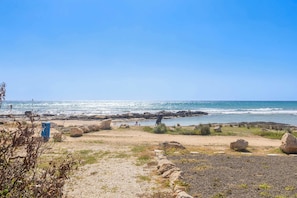 Ein Strand mit felsigem Gelände, wolkenlosem Himmel und dem Meer im Hintergrund.