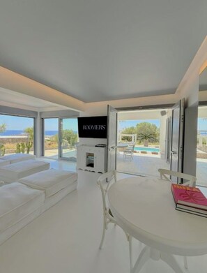 Un salón moderno con televisor, mobiliario blanco y vistas a una piscina y paisaje.