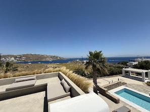 A poolside area with a view of the sea, a palm tree, and a modern building. - Casa Gekko Mykonos - Villa w Private Pool & Sea View (Mykonos)