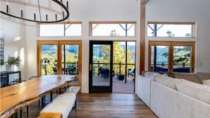 Ein geräumiger Raum mit einem Holztisch, Bänken, einer Couch und großen Fenstern mit Blick auf eine bewaldete Landschaft.