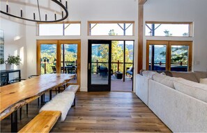 Ein geräumiger Raum mit einem Holztisch, Bänken, einer Couch und großen Fenstern mit Blick auf eine bewaldete Landschaft.