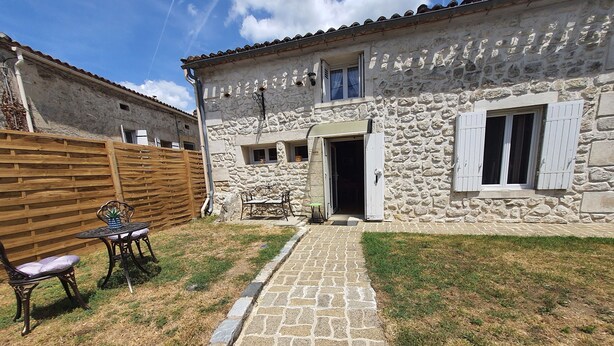 A stone building with a paved pathway leading to a door, a wooden fence, and outdoor furniture.