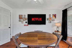 A dining area with a round wooden table, chairs, a television mounted on the wall, and framed artwork.