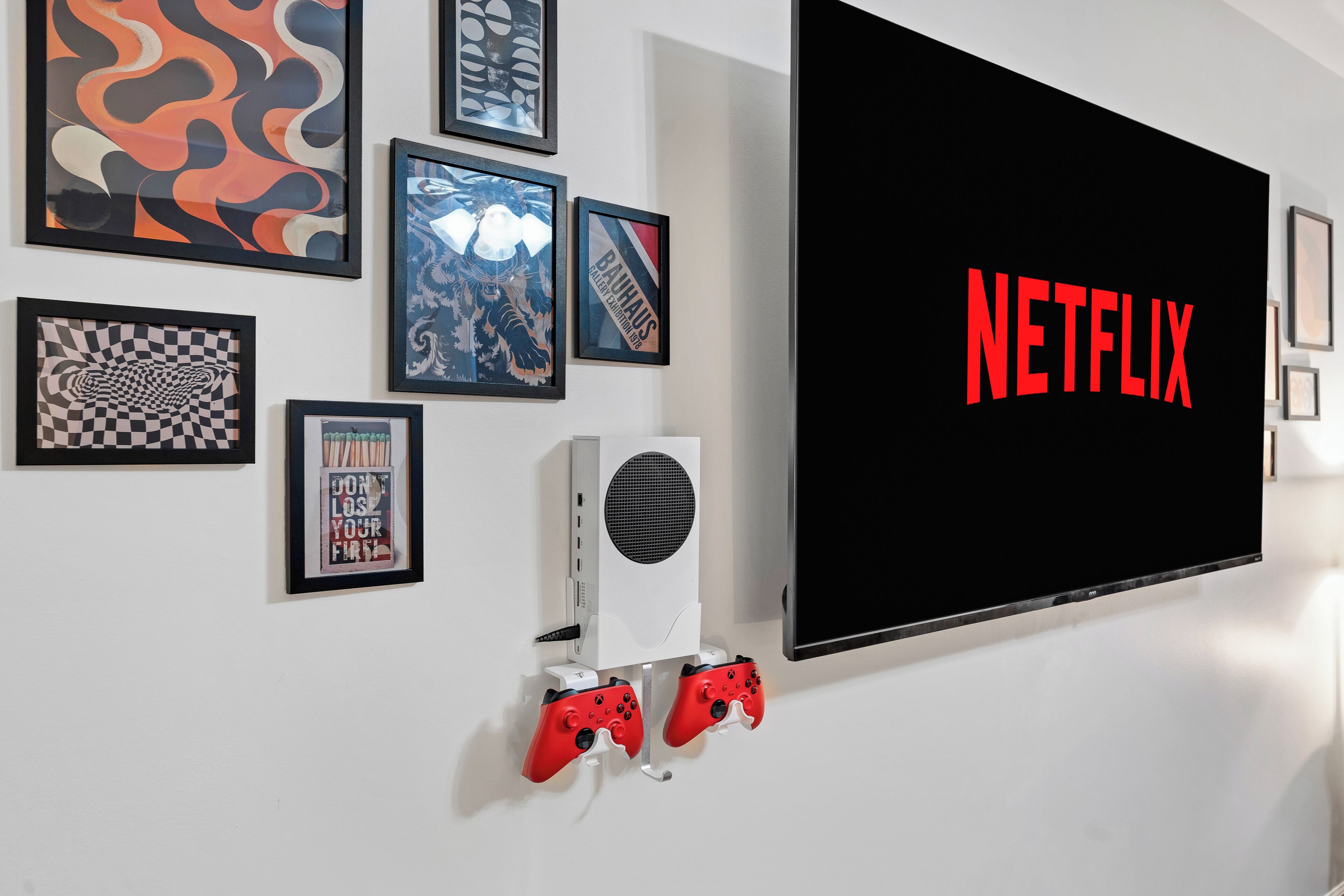 En væg med et stort fjernsyn, der viser Netflix-logoet, indrammet kunst og to røde spillekonsoller.