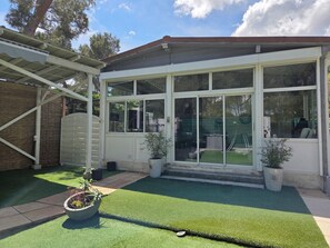 Ein überdachter Patio-Bereich mit Kunstgrasanlage, Topfpflanzen und Blick auf den Garten.
