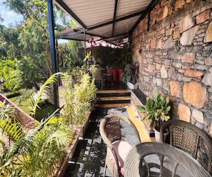 A covered outdoor area with stone walls, potted plants, and wicker furniture. - 1bhk Nature View Villa With Private Balcony (Udaipur)