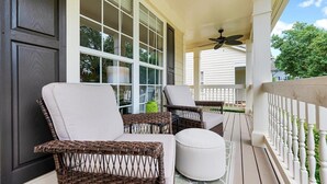 Eine überdachte Veranda mit Korbstühlen, einem weißen Hocker, großen Fenstern und einem Deckenventilator.