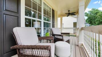 A covered porch with wicker chairs and a white ottoman, large windows, and a ceiling fan.
