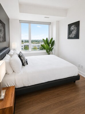 Ein modernes Schlafzimmer mit einem großen Bett, einem Nachttisch, einem Fenster mit Blick auf die Stadt und einem gerahmten Kunstwerk an der Wand.