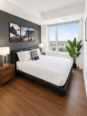 A modern bedroom with a large bed, a nightstand, a bedside table, a window with a view, and a potted plant.