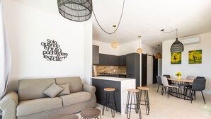 A modern living room with a sofa, coffee table, and bar stools. A kitchen area with a bar and dining table is visible in the background. - Symphony Sound By BarbarHouse (Sannicola)