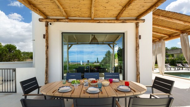 A wooden table set for six with chairs, a glass door leading to a pool area, and a view of the landscape. - Symphony Sound By BarbarHouse (Sannicola)