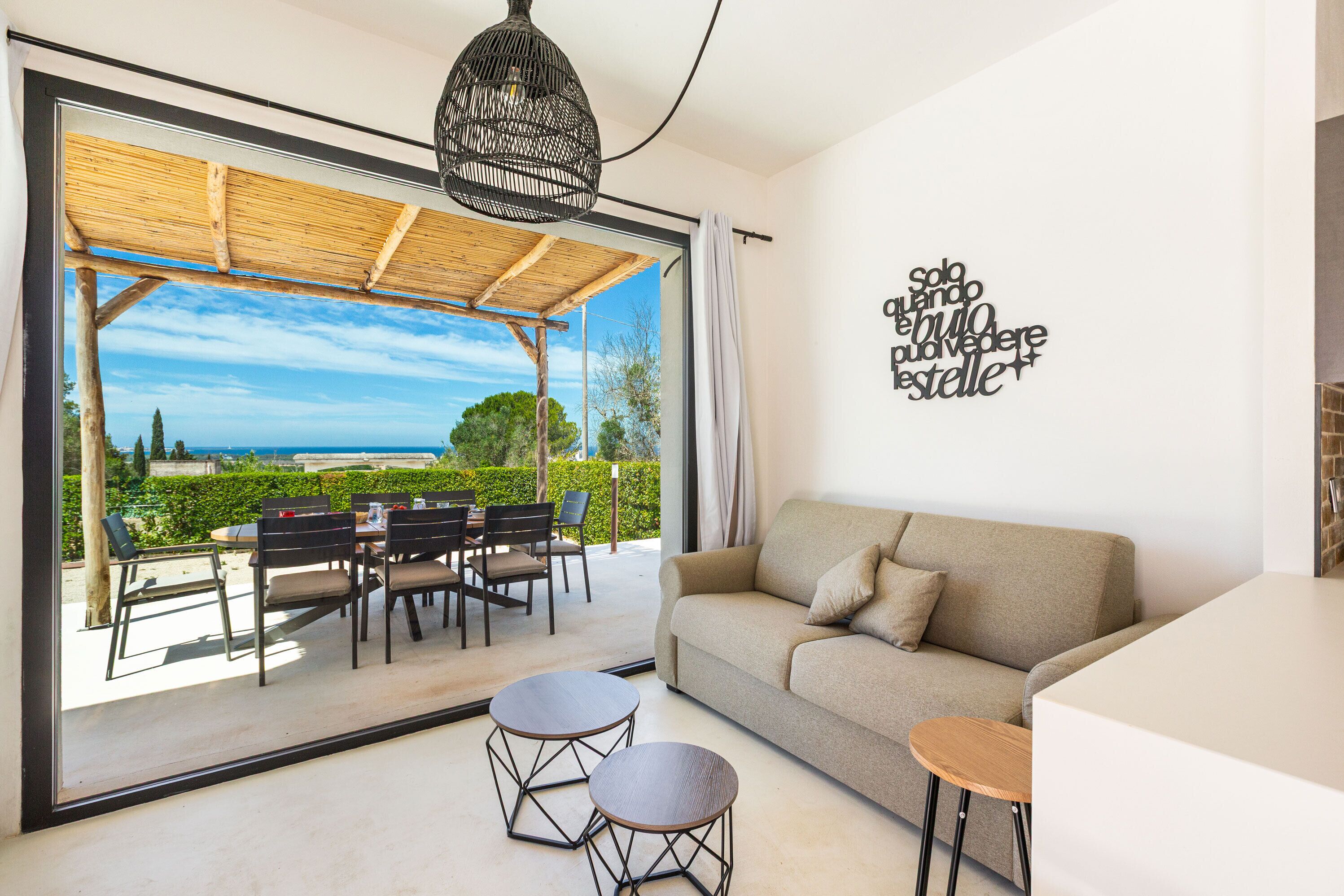 A modern living room with a sofa, two small tables, a dining area with chairs, and a large glass door leading to an outdoor patio with a view.