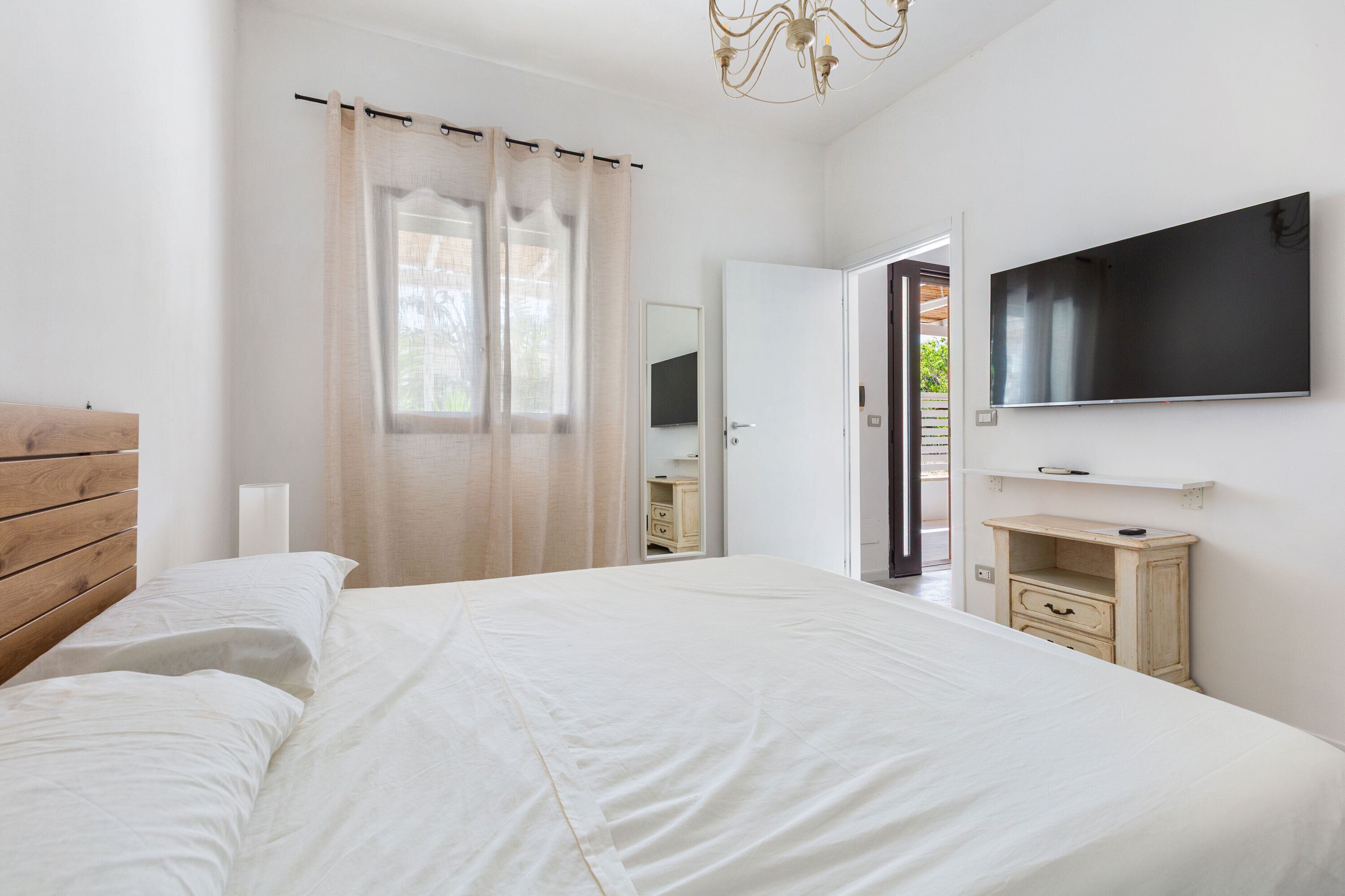 A bedroom with a bed, a TV mounted on the wall, a wooden headboard, a bedside table, and a window with curtains.
