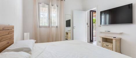 A bedroom with a bed, a TV mounted on the wall, a wooden headboard, a bedside table, and a window with curtains.